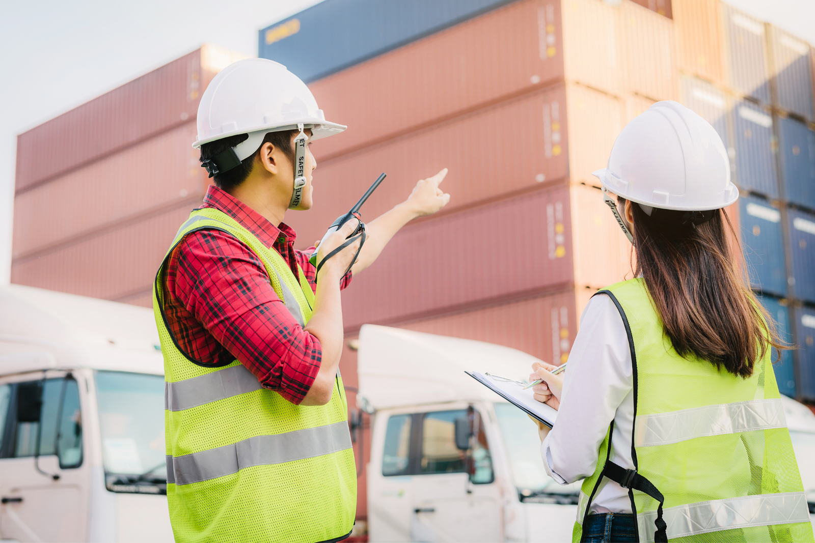 Two inspectors in hard hats and safety vests auditing stacked shipping containers before dispatch